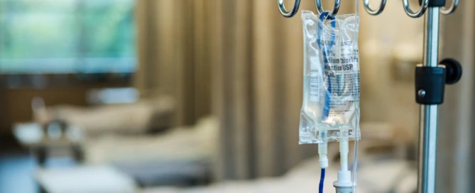 The focus of the photo is on the IV bag and stand in the foreground with empty hospital beds in a row in the background.